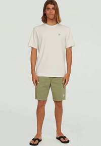 T-shirt crema a maniche corte con piccolo logo sul petto, abbinato a pantaloni corti verde oliva con una toppa, completato da ciabatte nere.