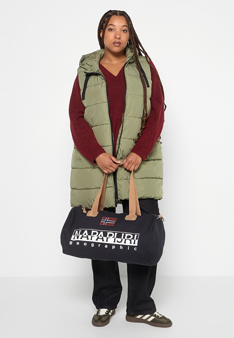 Vrouw met gevlochten haar in een groene puffer vest, bordeauxkleurige trui, zwarte jeans en sneakers, die een zwarte Napapijri reistas vasthoudt.