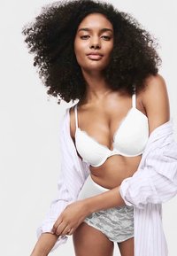 Reggiseno in pizzo bianco con bordi smerlati, mutandine in pizzo a vita alta abbinate e una camicetta leggera a righe, indossata sciolta sopra l'outfit.