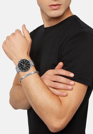 Montre en métal argenté avec un cadran noir, fonctionnalités de chronographe et un bracelet argenté, portée sur un bras nu aux côtés d'un bracelet tressé gris.