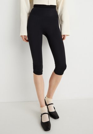 Fekete, fekvő anyagból készült capri leggings, magas derékpánttal és ráncolt részlettel a szegélyen, fekete cipővel és krémszínű zoknival párosítva.