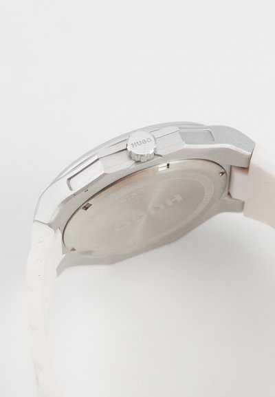 Montre en argent avec un boîtier texturé, bracelet en caoutchouc blanc, et couronne circulaire portant le nom de la marque "Hugo". Le dos a une finition lisse.