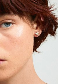 Silver flower stud earring with five petals and a central gem, worn on the ear. Short, textured hair and light skin are visible.