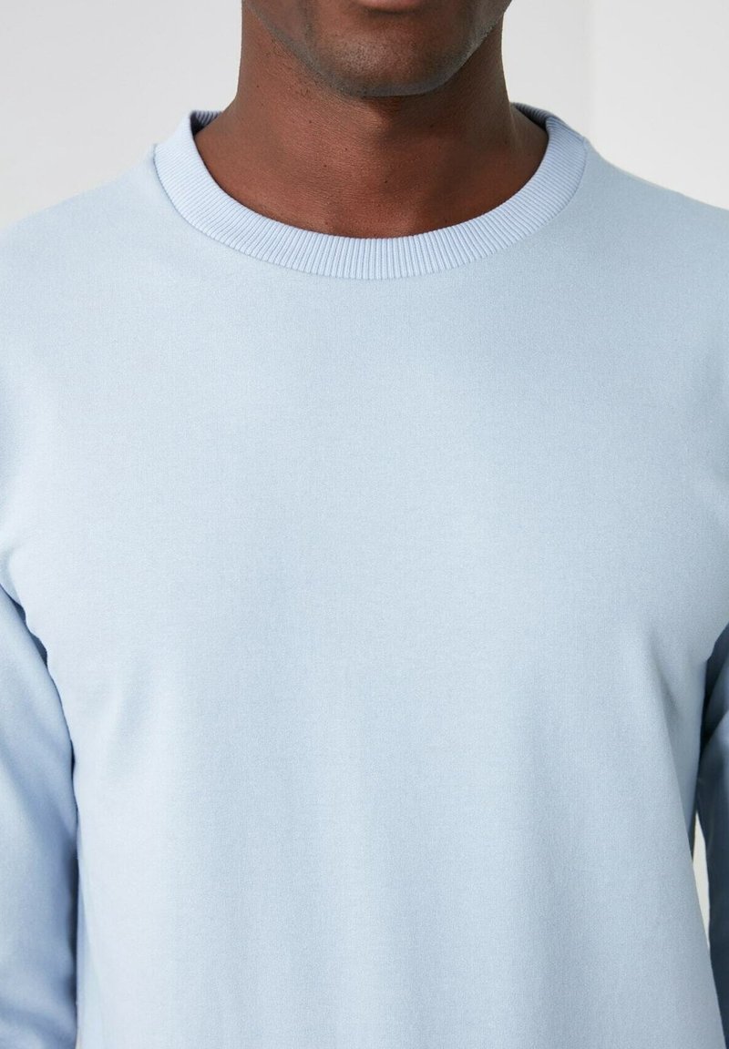 Chemise bleu clair à manches longues avec un col rond côtelé, confectionnée dans un tissu lisse à la texture douce. Aucun motif ou accent visible.