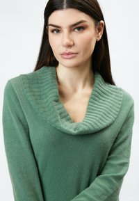 Grüner Pullover mit einem gerippten, oversized Kragen. Der Stoff wirkt weich und strukturiert, mit langen Ärmeln und einem lässigen Schnitt.
