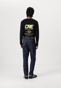 Black long-sleeve shirt with yellow graphic text on the back, paired with dark blue denim jeans and black shoes with white accents.