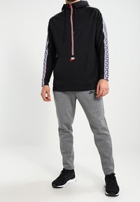 Herr svart hoodie med en framficka, röd dragkedja och mönstrade blåvita sidoränder. Matchas med grå mjukisbyxor och svarta skor.