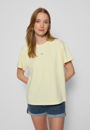 Vrouw met lang lichtbruin haar, die een gele oversized t-shirt en opgerolde spijkershort draagt, staand tegen een effen lichtgrijze muur.