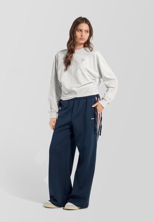Joven mujer con camiseta gris claro de manga larga y pantalones anchos azul marino con zapatillas cómodas, de pie frente a un fondo claro y liso.