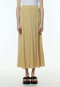 Geel, middellange rok gemaakt van textuurstof met een knoopsluiting aan de voorkant. Gepaard met dikke zwarte sandalen.
