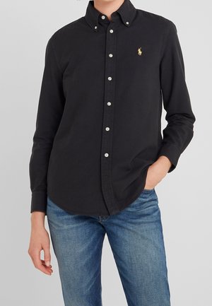 Persoon draagt een zwart overhemd met knopen met een klein geel logo op de borst en blauwe spijkerbroek, één hand in de zak tegen een effen achtergrond.