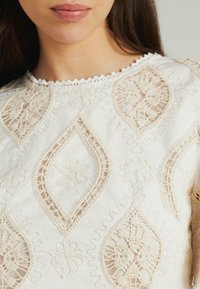 Witte katoenen top met gedetailleerde beige borduursels in bloem- en geometrische patronen, met een golvende halslijn en een gestructureerde afwerking.