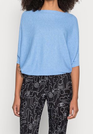 Jasnoniebieski, prążkowany, oversized sweter z szerokim dekoltem, w połączeniu z czarnymi spodniami z białymi abstrakcyjnymi wzorami i detalami w postaci pasków.