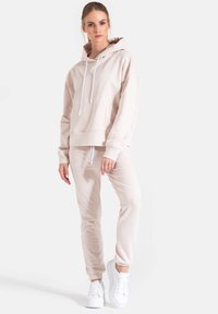 Jasno beżowy sweatshirt z kapturem i sznurkiem, w zestawie z dopasowanymi dżinsami dresowymi. Białe sneakersy dopełniają stylizację. Gładka tekstura.