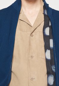 Mörkblå zip-up jacka över en beige mockaskjorta med bröstficka, med ett mönstrat mörkt tyg under, som visar distinkta färgkontraster.