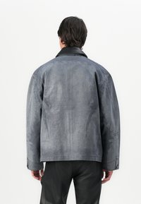 Άνδρας φορεμένος oversized γκρι σακάκι με μαύρο γιακά και μαύρο παντελόνι, στέκεται μπροστά σε λευκό τοίχο.