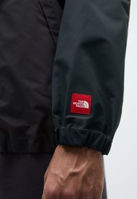 Svart och grön jackärm med elastiskt manschett, med en framträdande röd fyrkantig logotyp av The North Face. Slätt, tekniskt tyg.