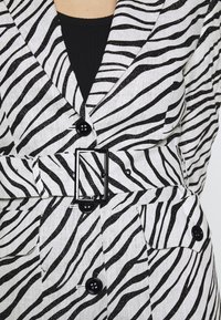 Gros plan d'un blazer à rayures zébrées noires et blanches avec des boutons noirs, une ceinture assortie avec boucle carrée, et un top intérieur noir.