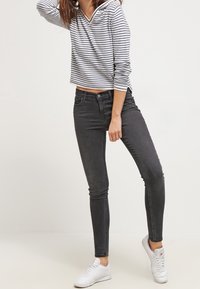 Donna che indossa jeans skinny neri, sneakers bianche e una maglietta bianca a maniche lunghe con strisce orizzontali nere, in piedi contro uno sfondo neutro.