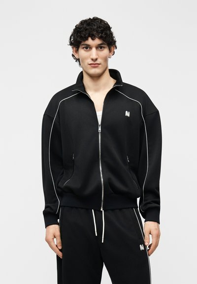 Jeune homme aux cheveux bouclés portant une veste noire zippée et un pantalon noir assorti avec des liserés blancs et un logo, debout devant un fond uni.
