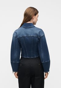 Γυναίκα που φοράει κοντό, μπλε τζιν μπουφάν δύο τόνων με μακριά μανίκια και μαύρο παντελόνι, από πίσω σε απλό φόντο.