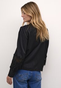 Blusa negra con detalles de encaje en las mangas, que presenta un ajuste relajado y mangas largas con puños. Combinada con jeans de mezclilla azul.