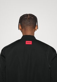 Veste bomber noire avec un écusson rouge portant le texte "HUGO" dans le dos, texture lisse, col côtelé. Vue arrière avec cheveux tressés visibles.