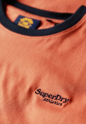 Camiseta de algodón melocotón con cuello acanalado en navy y logo bordado "SuperDry Athletics" en el pecho. Textura suave.