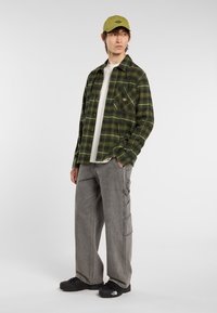 Een jonge man staat casual, hij draagt een groen geruit overhemd over een witte t-shirt, grijze cargo broek en zwarte schoenen, met een groene pet.