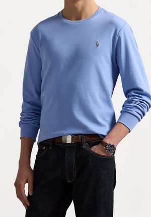 Mann trägt hellblaues Langarmhemd mit kleinem besticktem Logo, dunkle Jeans, braunen Gürtel und Armbanduhr am linken Handgelenk.