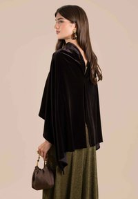 Capa de terciopelo negro con un diseño drapeado, que presenta un escote en V y un acabado texturizado, combinada con una falda verde metálica y un bolso de mano marrón.