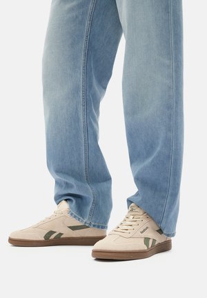Jasnoniebieskie dżinsy zakrywające beżowe i oliwkowe buty sportowe Reebok z brązowymi podeszwami na gładkim jasnym tle.