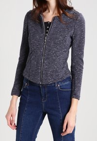 Donkerblauwe jas met ritssluiting met licht gevlekt patroon, lange mouwen, gestructureerde stof en een getailleerde pasvorm, gecombineerd met donkerblauwe spijkerbroeken.