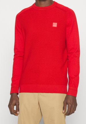 Homme portant un pull texturé rouge vif à manches longues avec un petit patch logo sur la poitrine, associé à un pantalon beige.