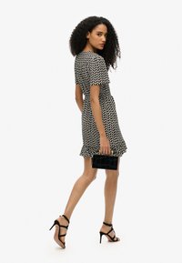 Vestido corto de estampado en blanco y negro con dobladillo rizado, complementado con un bolso de mano de charol negro y sandalias de tacón alto.