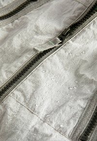 Vesiä kestävä harmaa kangas, jossa on näkyviä vesipisaroita. Varustettu mustilla vetoketjuilla. Sileä pintatekstuuri, kevyt ja kestävä muotoilu.