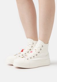 Zapatillas de lona blancas altas con suelas de goma, que cuentan con encantos coloridos en los cordones y una puntera texturizada.