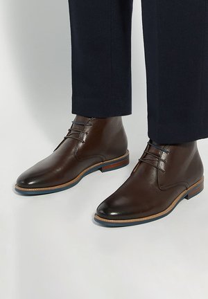 Braune Leder-Stiefeletten mit Schnürsenkeln, getragen zu dunkelblauen Hosen vor schlicht hellem Hintergrund.