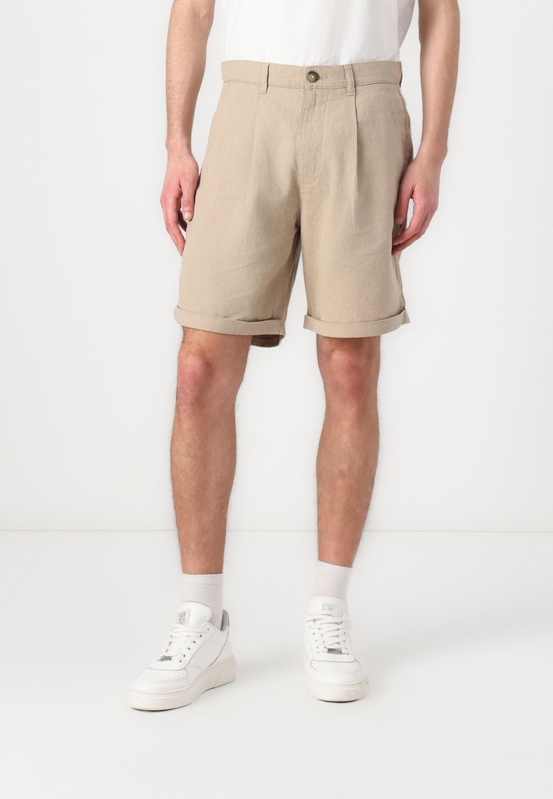 Bež ljetne hlače od lana s prevrnutim rubovima, prednjim nabranim detaljima i jednim dugmetom, u kombinaciji s bijelim tenisicama i čarapama do gležnja.