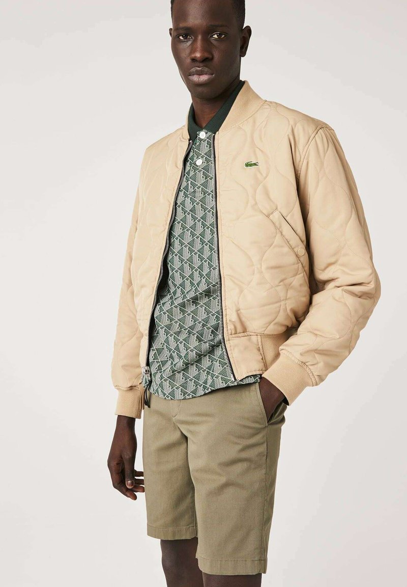 Veste bomber matelassée beige, chemise à motif vert, shorts khaki. Comprend des poignets et un col côtelés, avec un détail de logo sur la veste.