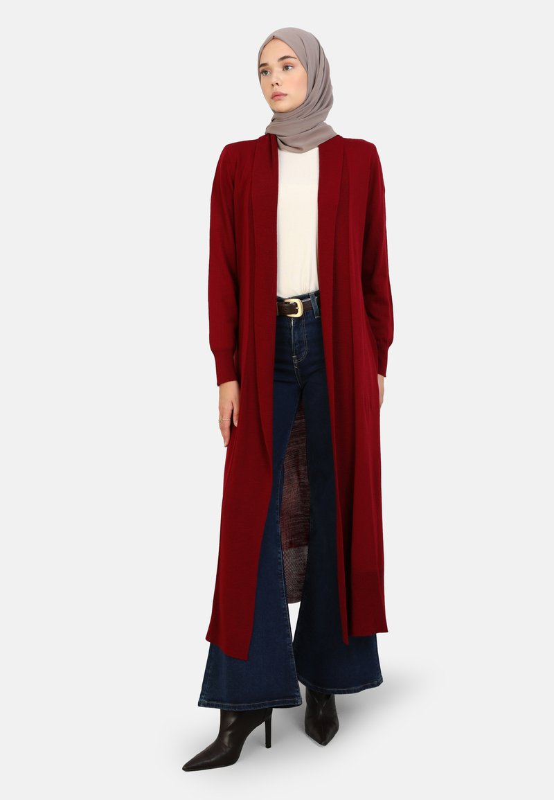 Long, open-front cardigan in deep red knit fabric, worn over a white top and blue flared jeans, paired with black high-heeled boots.