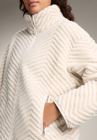 Cremfarbene Fleecejacke mit einem Zickzackmuster, hohem Kragen, Reißverschlusstaschen und einem glatten Reißverschluss. Weicher Stoff mit lockerer Passform.