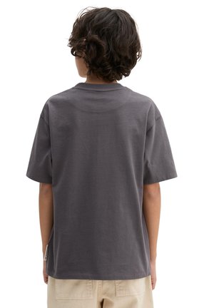 Enfant aux cheveux bouclés foncés portant un t-shirt ample manches courtes gris foncé et un pantalon beige clair, debout de dos sur un fond blanc.