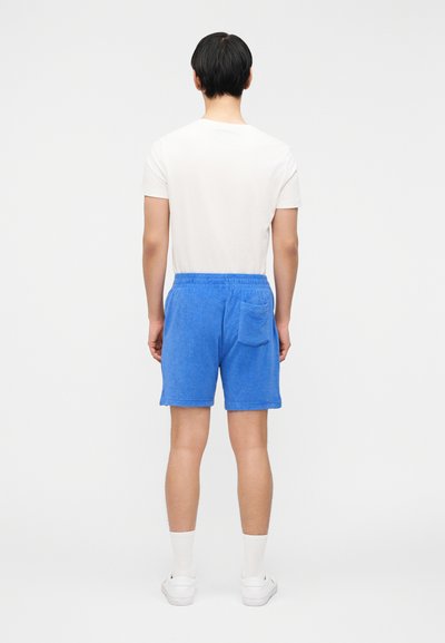 Άνδρας με πλάτη φοράει λευκό πουκάμισο με κοντά μανίκια, φωτεινά μπλε σορτς από πετσέτα με πίσω τσέπη, λευκές κάλτσες και λευκά αθλητικά, στέκεται σε απλό φόντο.