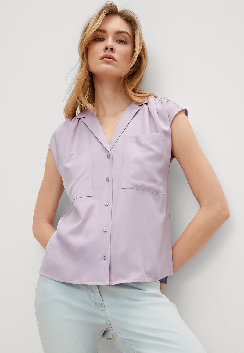 Lavender short-sleeve shirt with a notched collar, two front pockets, and button closure. Smooth fabric with a subtle sheen.