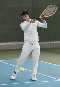 Veste de sport blanche et pantalon, joueur balançant une raquette de tennis avec une poignée orange, arrière-plan vert, court bleu, balle de tennis jaune visible.