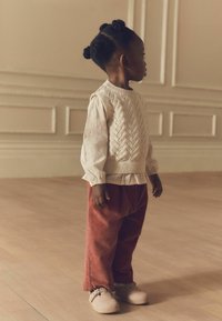 Strikket sweater med zigzag-mønster, hvid langærmet bluse, rustfarvede fløjl bukser og beige klip-klapper på et trægulv.