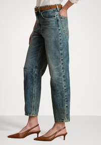Svetlo modri denim hlače s sproščenim krojem, ki imajo sprednji žep, vidno šivanje in svetlo rjav pas. Ujemajo se z rjavimi čevlji na peto.