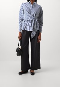 Camicia a righe blu e bianche con design avvolgente, abbinata a pantaloni larghi scuri e ballerine nere; tiene una piccola borsa nera.