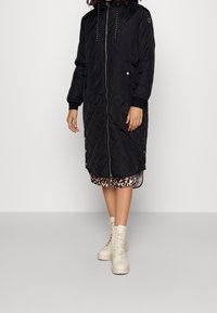 Abrigo largo acolchado negro con capucha, cierre de cremallera y detalles a presión en los laterales. Llevado sobre un vestido de estampado de leopardo y combinado con botas blancas de cordones.